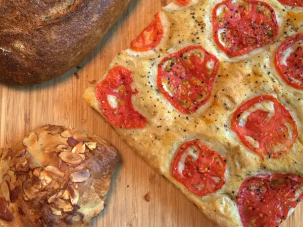 Focaccia receta con tomates frescos y hierbas, junto a un pan rústico y un bollo con almendras.