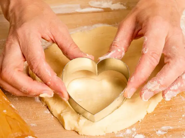 Manos cortan masa con un cortador en forma de corazón, parte de una receta de galletas.