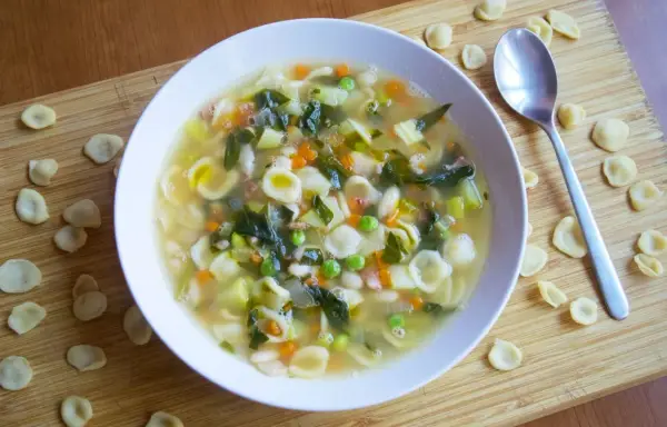 Un reconfortante plato de sopa minestrone, lleno de pasta orecchiette, verduras y frijoles, servido en un tazón blanco sobre una tabla de madera.