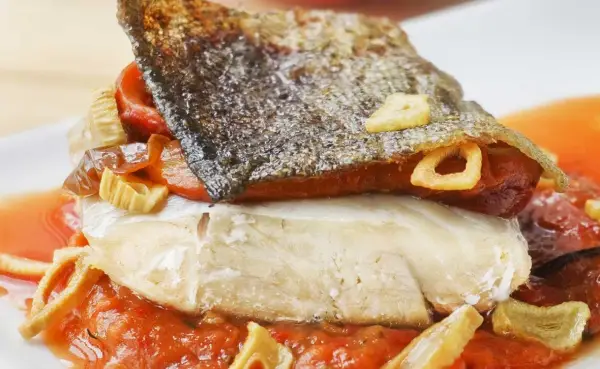 Bacalao a la riojana con piel crujiente, pimientos y ajo frito, servido sobre una cama de salsa de tomate.